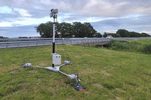 Op de foto staat langs een snelweg een controleapparaat dat verplaatst kan worden. Het apparaat bestaat uit een metalen statief met daarop op een paal met bovenaan een camera of sensor. Het apparaat staat in de berm naast de vangrail.