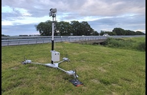 Op de foto staat langs een snelweg een controleapparaat dat verplaatst kan worden. Het apparaat bestaat uit een metalen statief met daarop op een paal met bovenaan een camera of sensor. Het apparaat staat in de berm naast de vangrail.