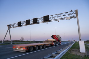 Op de foto rijdt een vrachtwagen onder een metalen bouwwerk (poort) door boven de snelweg. Aan het bouwwerk hangen meerdere camera's of sensoren.