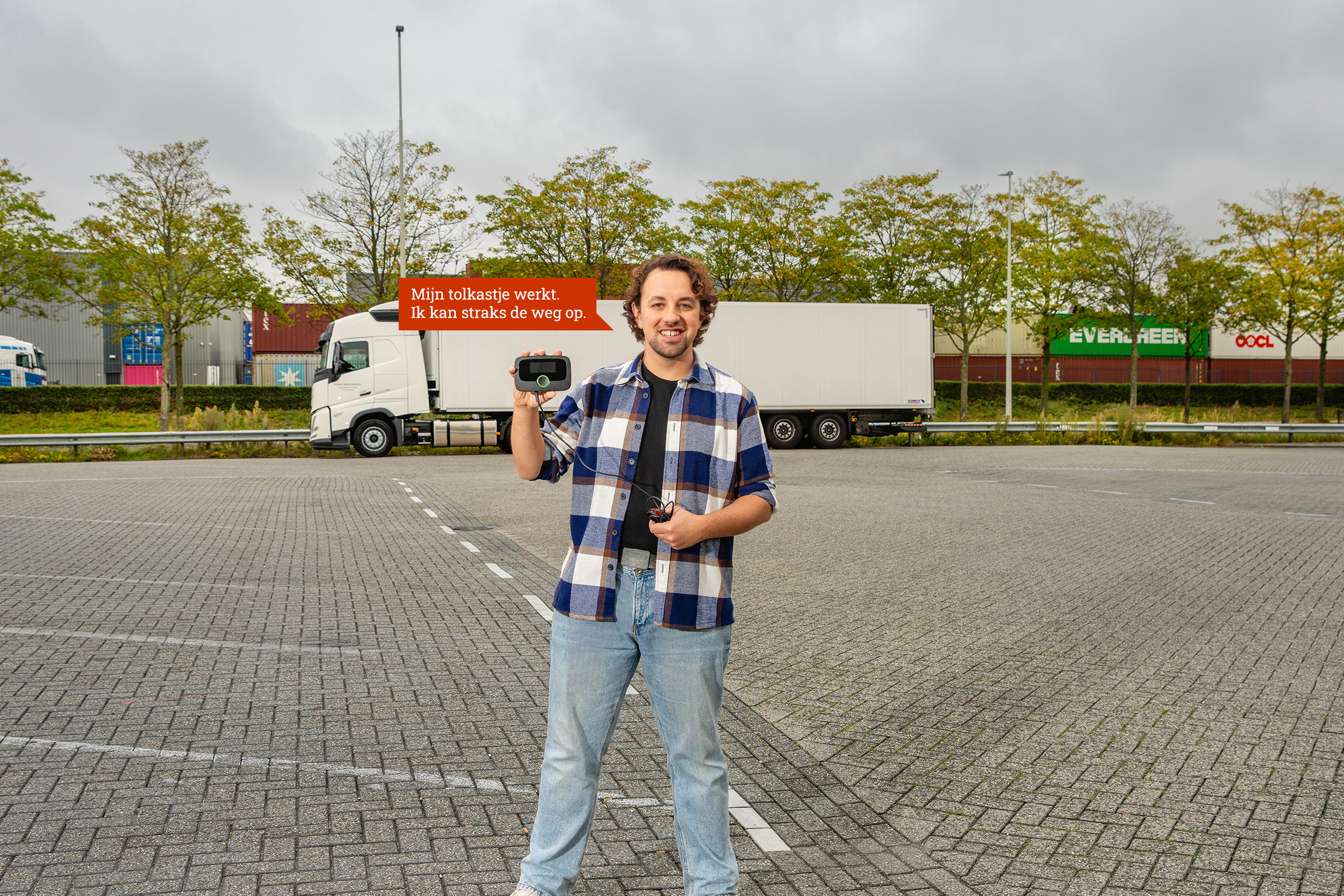 Een vrachtwagenchauffeur staat voor zijn vrachtwagen met een tolkastje in zijn hand. Het lampje van het kastje is groen. Naast de man is een tekstballon afgebeeld met de tekst: 'Mijn tolkastje werkt. Ik kan straks de weg op.'