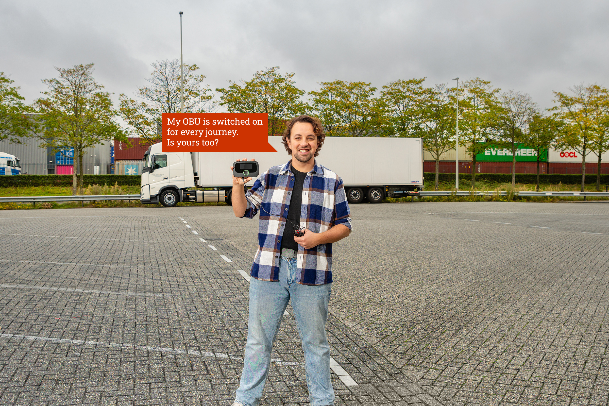 A lorry driver is standing in front of his lorry holding an OBU in his hand. The light on the OBU is green. Next to the man is a speech bubble containing the text: ‘My OBU is switched on for every journey. Is yours too?’'
