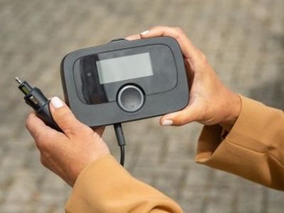 An OBU is held with one hand. The rectangular electronic device is roughly the size of a hand. In addition to a small display and a power button on the OBU, a cable with a plug can be seen. This plug fits into the 12- or 24-volt socket of a truck.